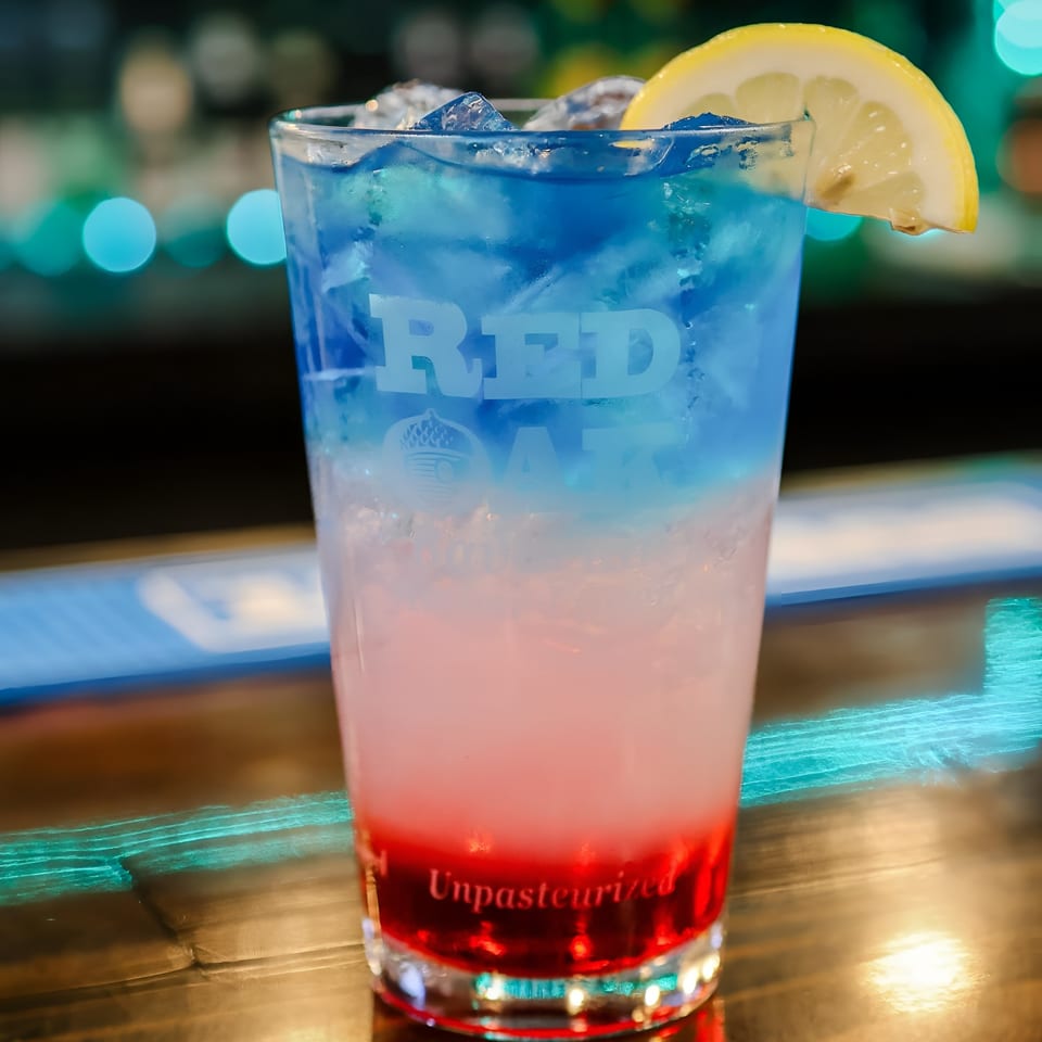 A Red Oak branded glass filled with a red, white, and blue layered drink, garnished with a lemon slice.