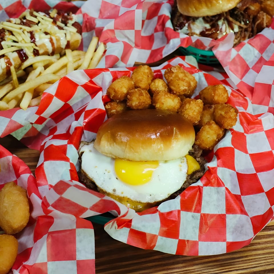 A burger with a fried egg and tater tots in a red and white checkered paper-lined basket.