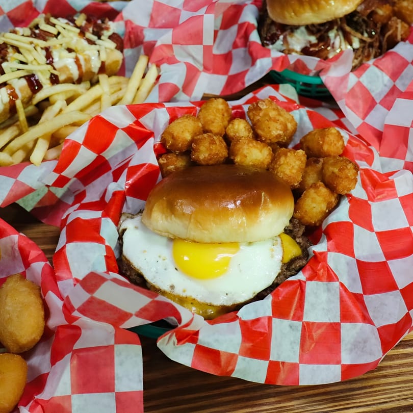 A burger with a fried egg and tater tots in a red and white checkered paper-lined basket.