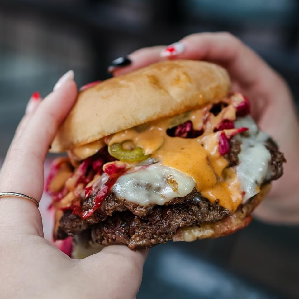 Hands holding a double smash burger with melted cheese, special sauce, pickled jalapeños, and red cabbage.