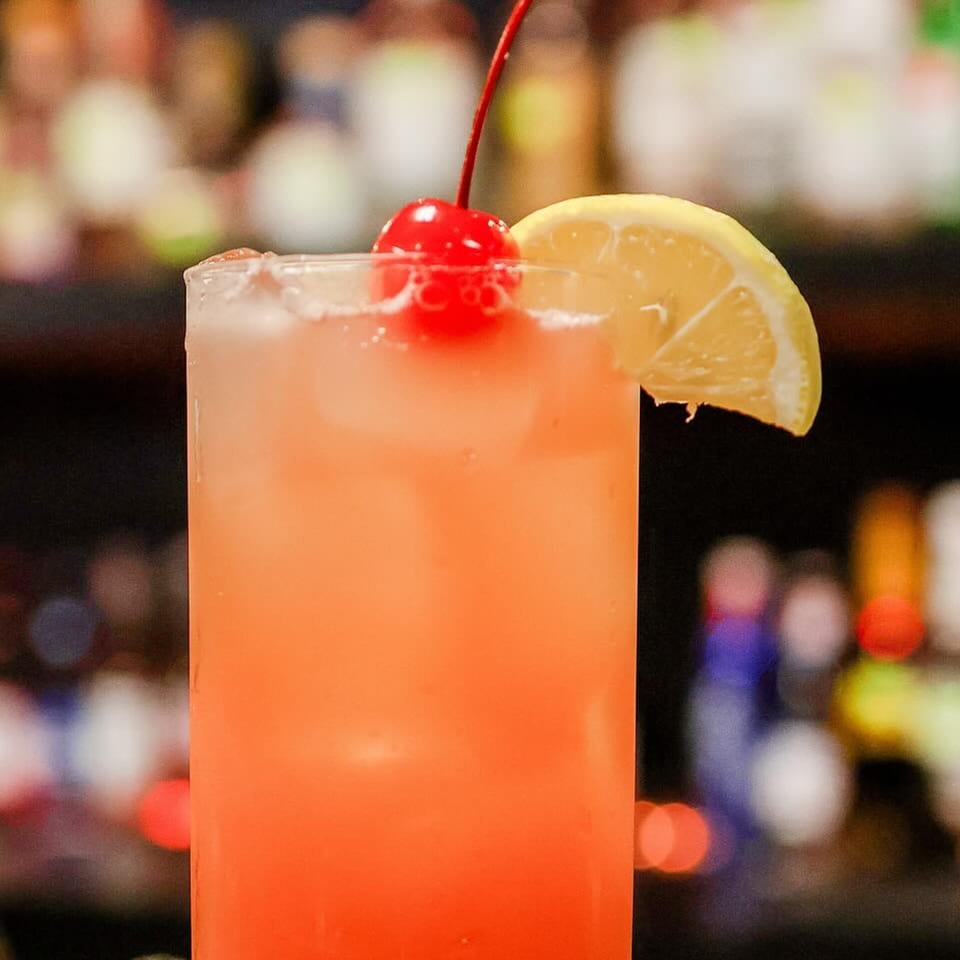 A vibrant orange cocktail with ice, garnished with a red cherry and a lemon slice.