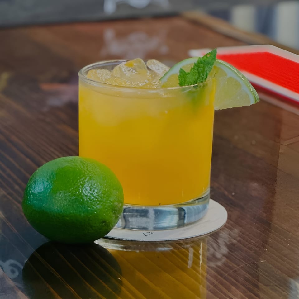 A vibrant yellow cocktail with ice, garnished with a lime wedge and mint, next to a whole lime on a wooden table.