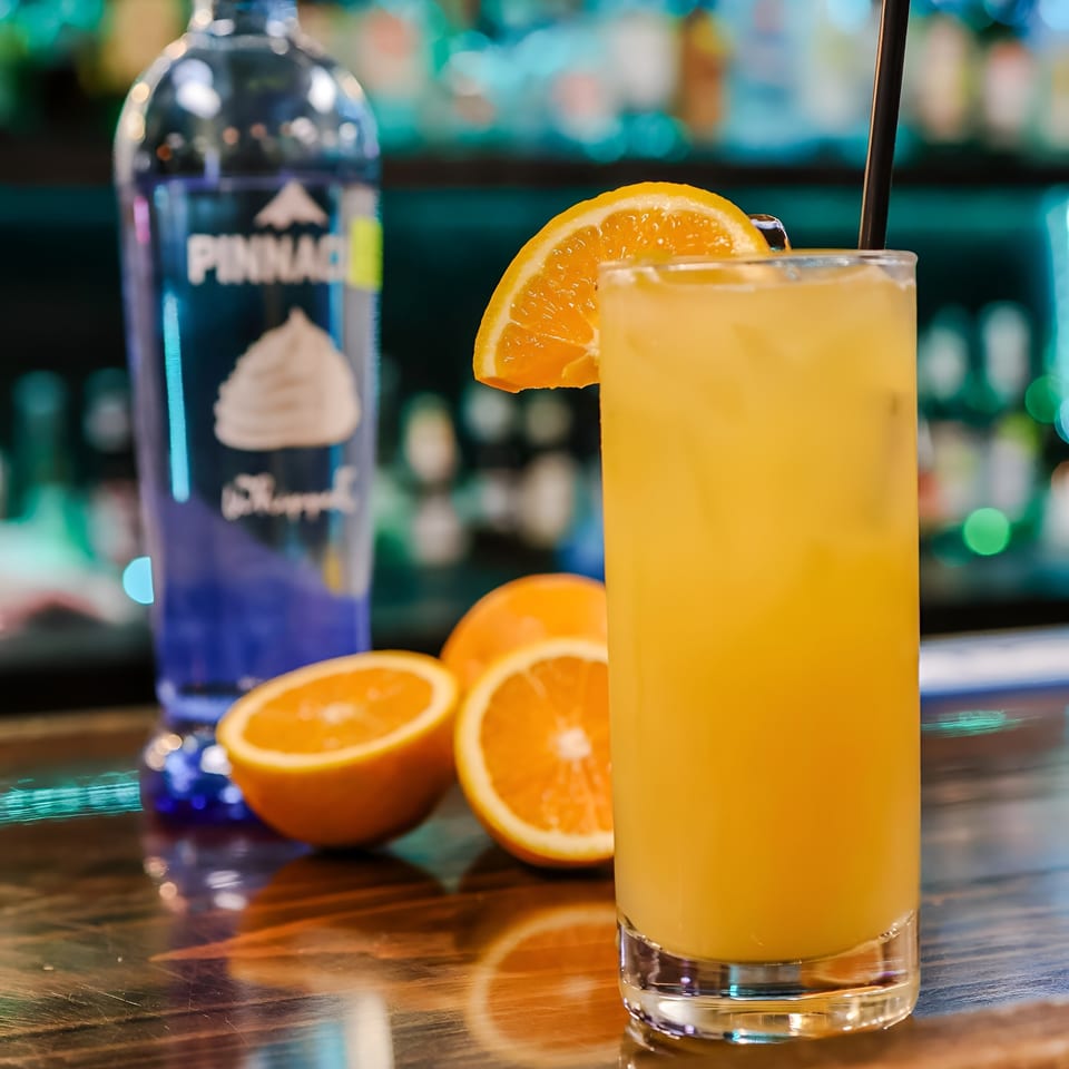 Orange cocktail with a slice of orange and a black straw, next to a bottle of Pinnacle vodka.