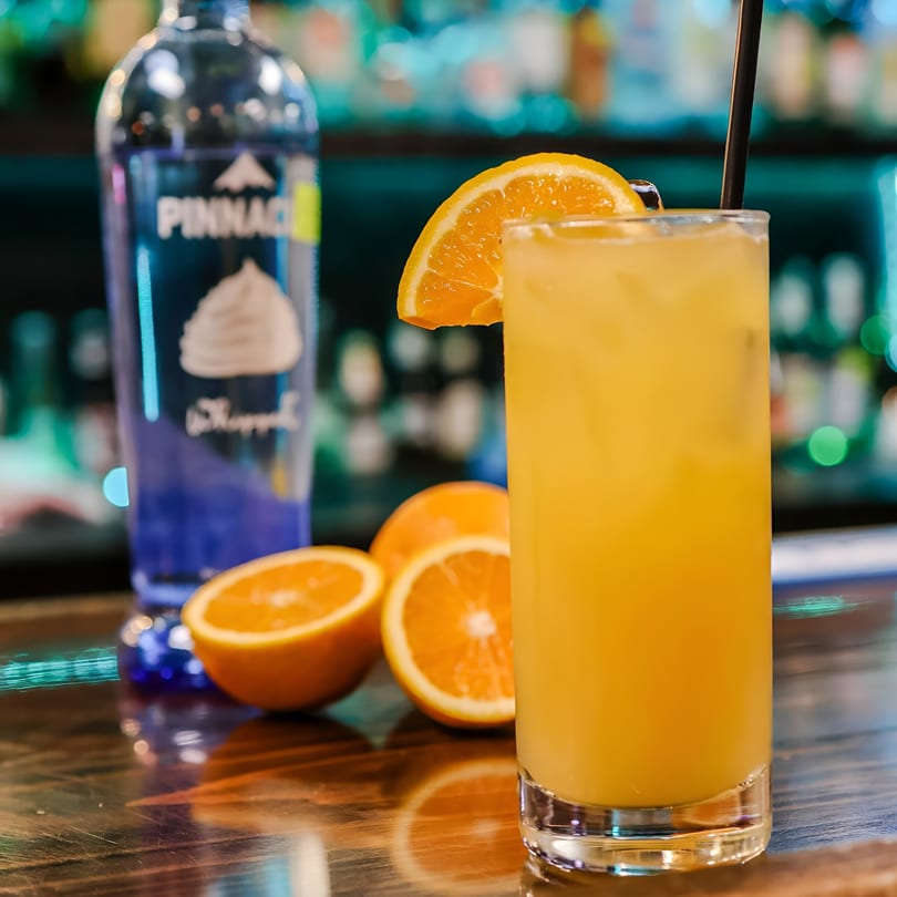 Orange cocktail with a slice of orange and a black straw, next to a bottle of Pinnacle vodka.