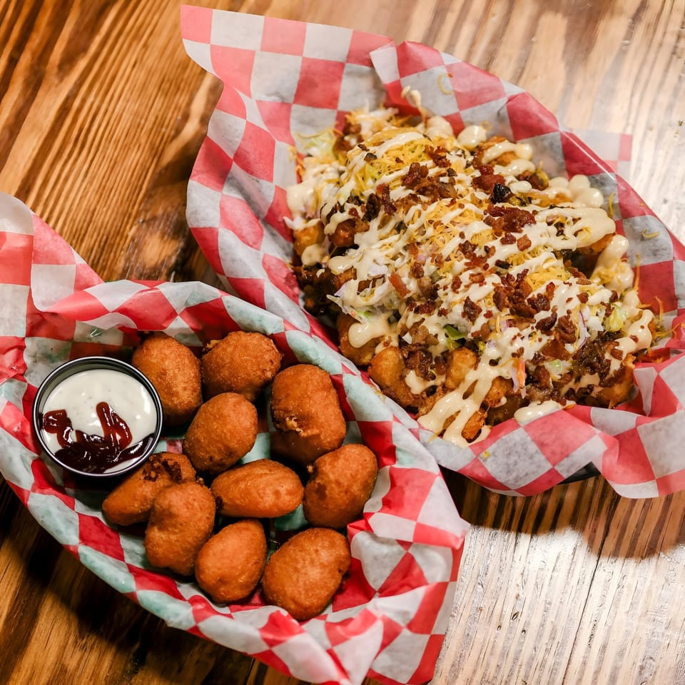 Two baskets of fried food on a wooden table: one with loaded tater tots, the other with hush puppies and dipping sauces.