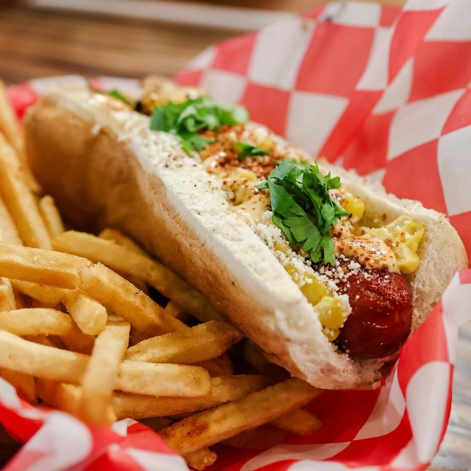Gourmet hot dog with corn, cheese, and cilantro, served with a side of fries in a red checkered basket.