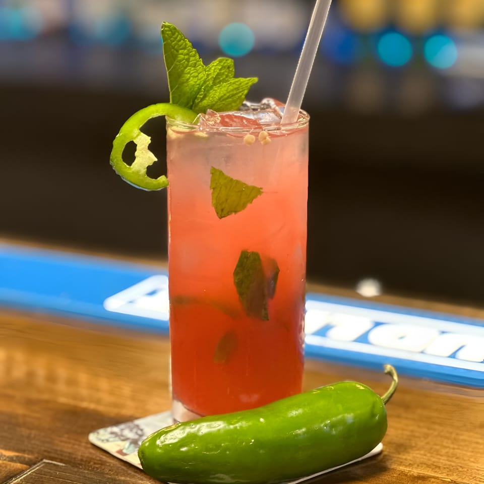 Pink cocktail with mint, a jalapeño garnish, and a whole jalapeño on a bar.