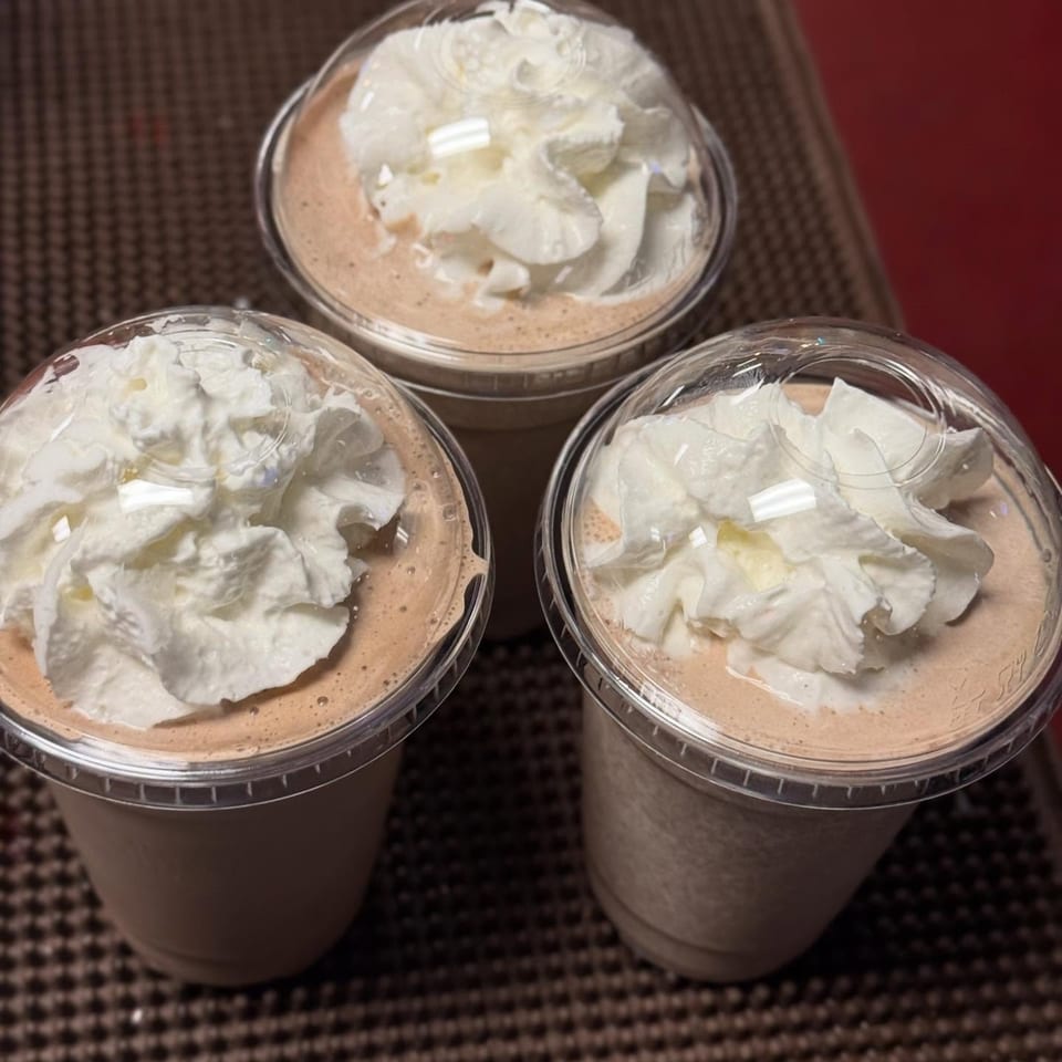 Three chocolate milkshakes with whipped cream and dome lids on a brown textured surface.